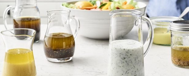 Woman making 7 homemade versatile pantry dressings with a fresh colorful salad on a white marble counter in a beautiful kitchen