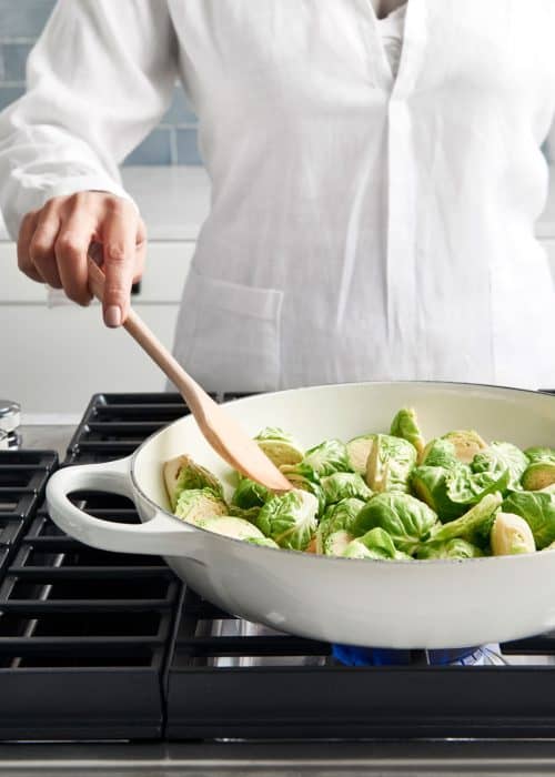 Woman in white shirt cooking candied brussels sprouts on a stove in a white skillet