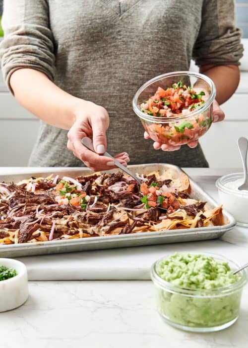 Woman putting pico de gallo on a sheetpan of steak nachos with guacamole on a white marble surface