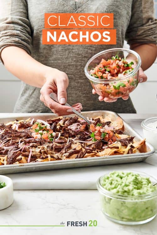 Woman putting pico de gallo on a sheetpan of steak nachos with guacamole on a white marble surface