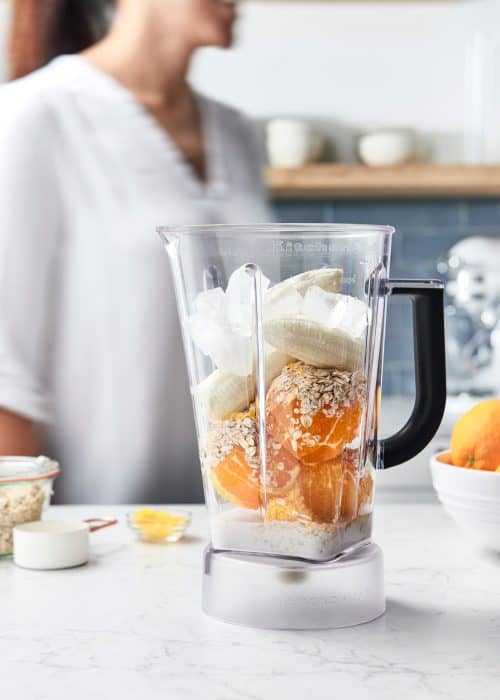 Woman making a smoothie with fresh oranges and bananas in a blender in a beautiful white marble kitchen