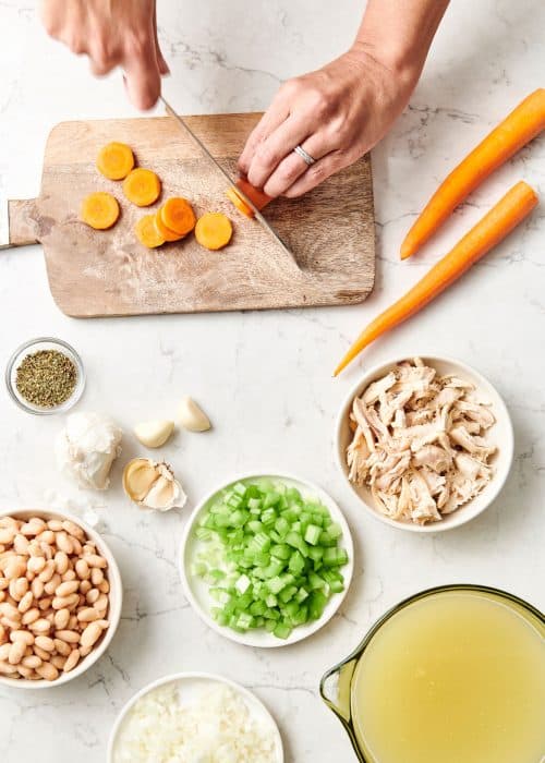 Woman cutting vegetables on a wood cutting board with ingredients to make chicken soup on a white marble countertop