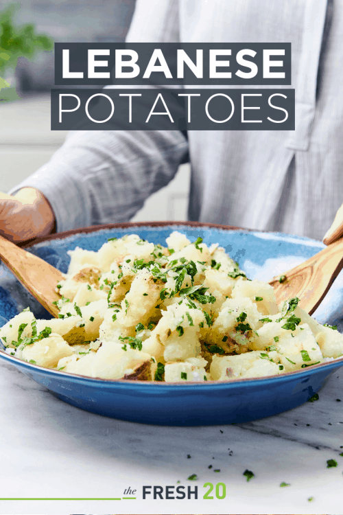 Woman tossing potatoes garnished with chives in a blue bowl with wooden spoons on a white marble surface