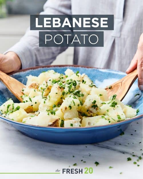 Woman tossing potatoes garnished with chives in a blue bowl with wooden spoons on a white marble surface