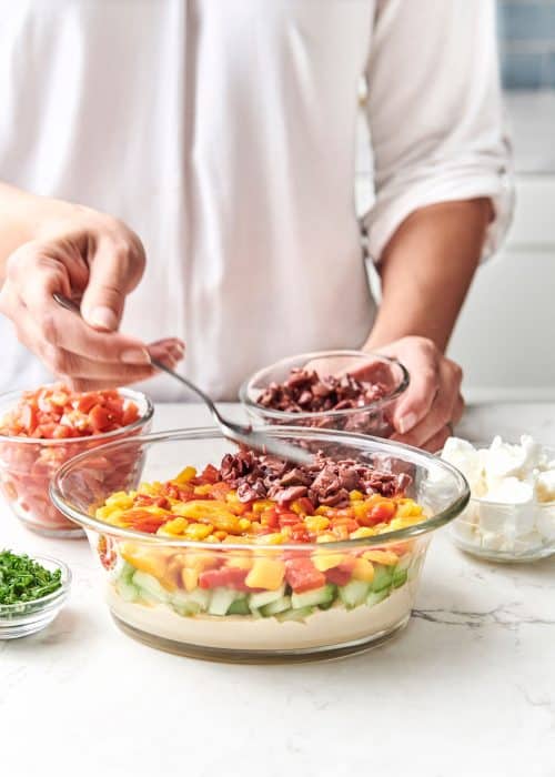 Woman using a spoon to layer olives into a serving bowl to create an easy 7-layer Mediterranean dip for entertaining