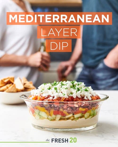 People conversing behind a vibrant glass bowl full of 7 layer dip garnished with parsley on a white marble surface next to pita chips