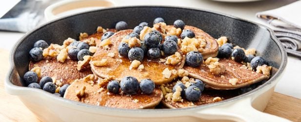 Skillet full of fluffy pancakes with plump blueberries, crushed nuts & syrup in a white marble kitchen