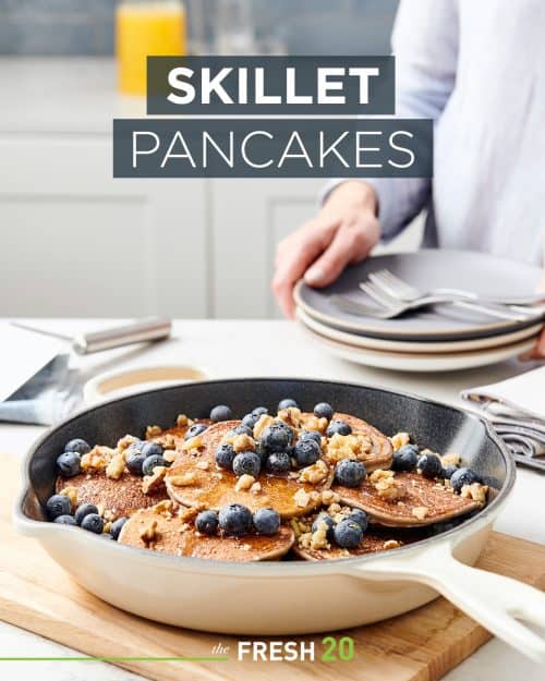 Skillet full of fluffy pancakes with plump blueberries, crushed nuts & syrup in a white marble kitchen