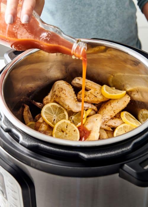 Pouring hot sauce into Instant Pot full of buffalo chicken wings & lemon slices