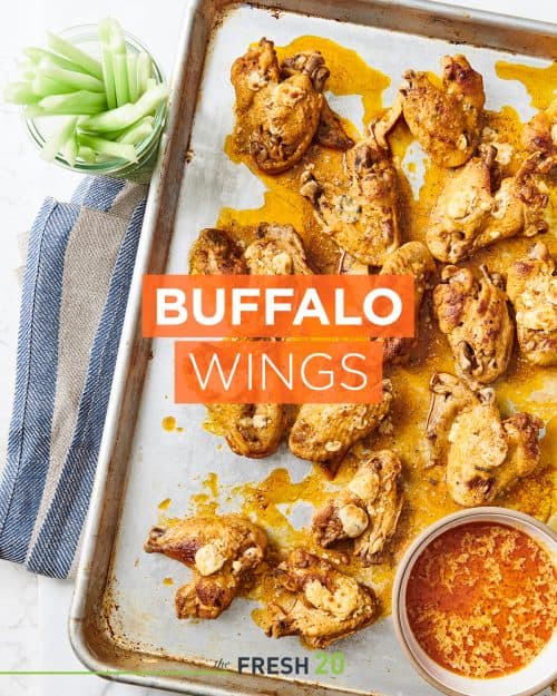 Baking sheet pan full of buffalo wings with a bowl of dipping sauce and celeryon a white marble surface