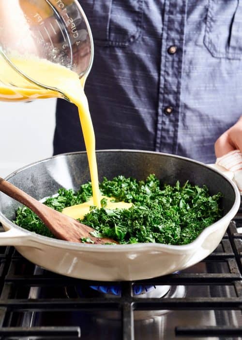 Man pouring egg mixture into a skillet full of kale cooking on gas cooktop