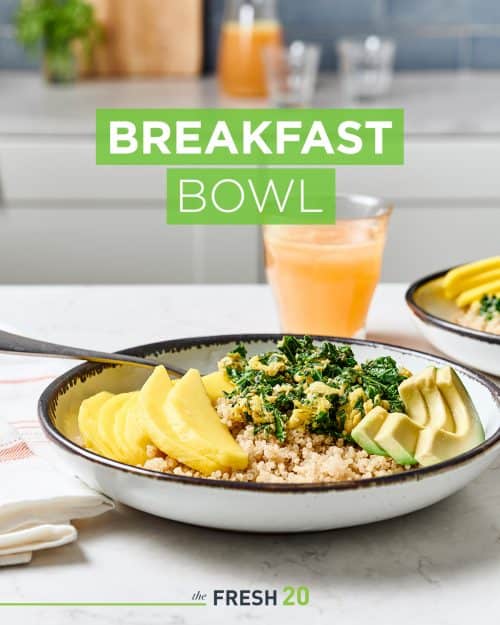 Kale, avocado, quinoa and sliced mango breakfast bowl with a glass of fresh juice on a white marble surface