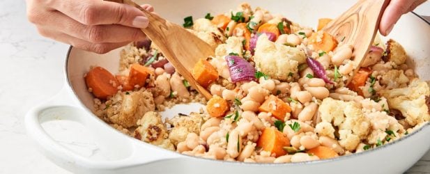 Tossing an easy roasted cauliflower cumin vegetable salad with two wooden spoons in a white skillet on a white marble counter