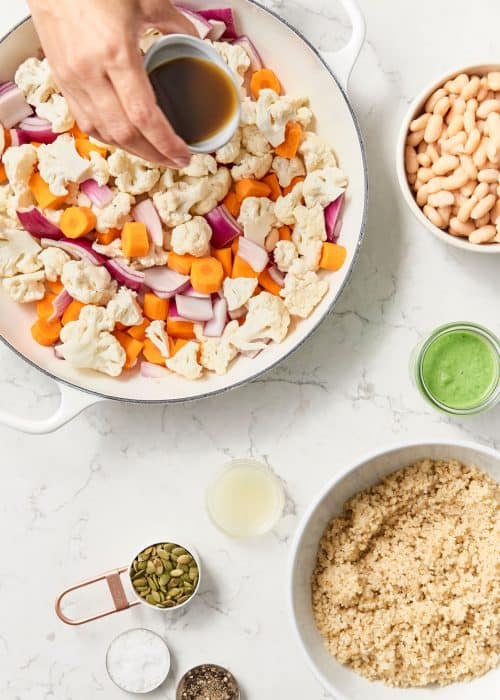 Hand pouring dressing on an easy cumin roasted vegetable salad in a white skillet with bowls of ingredients on a white marble counter