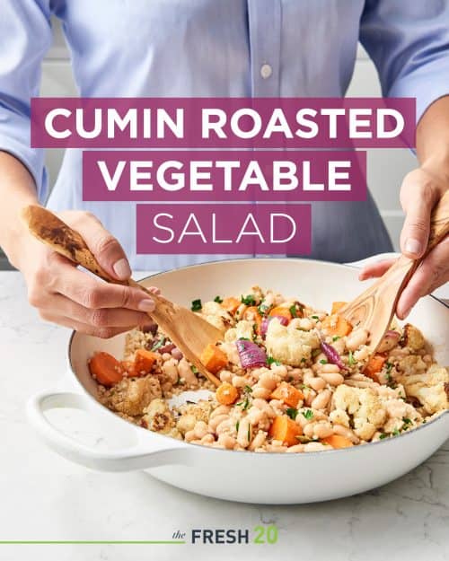 Woman tossing an easy roasted cauliflower cumin salad with two wooden spoons in a white marble kitchen