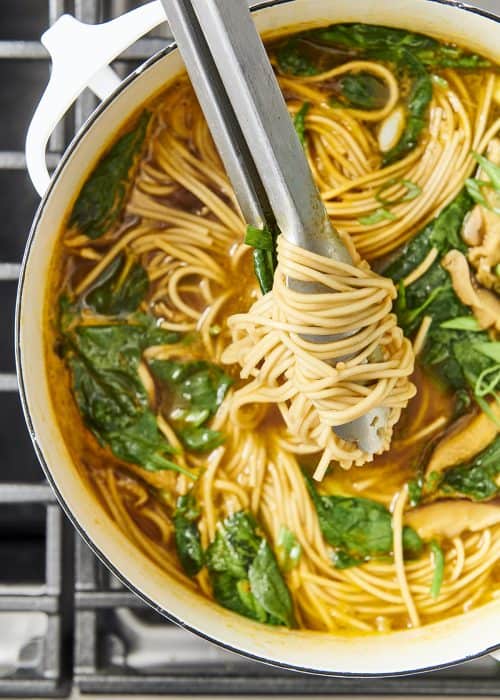 Silver tongs grabbing hearty ramen noodles from stockpot full of noodles, spinach and onions on a metal cooktop