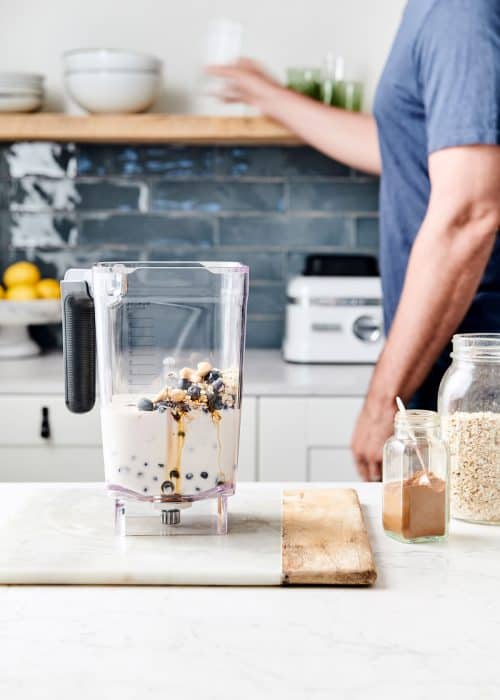 Man preparing a vegan smoothie in a blender full of creamy almond milk, honey, cinnamon and blueberries in a beautiful white marble kitchen