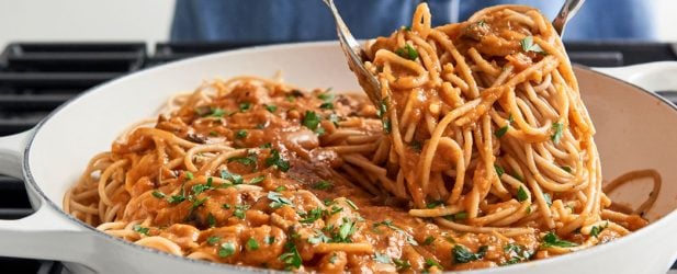 Scooping vegan bolognese with two white serving utensils from a skillet on a cooktop