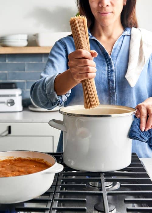 Woman placing uncooked pasta noodles into a cream colored pot next to pasta sauce on a metal stovetop