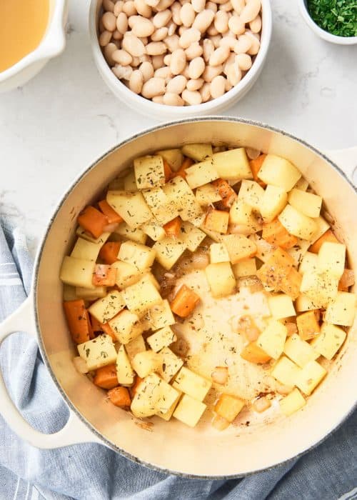 Le Cruset pot full of cubed potatoes and carrots alongside a bowl of white beans on a white marble surface