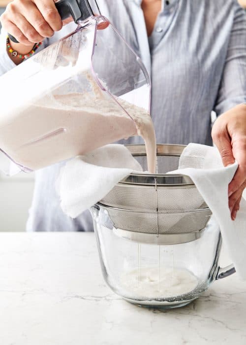 Woman pouring blended almonds into a strainer with cheese cloth to drain into a glass bowl in a white marble kitchen