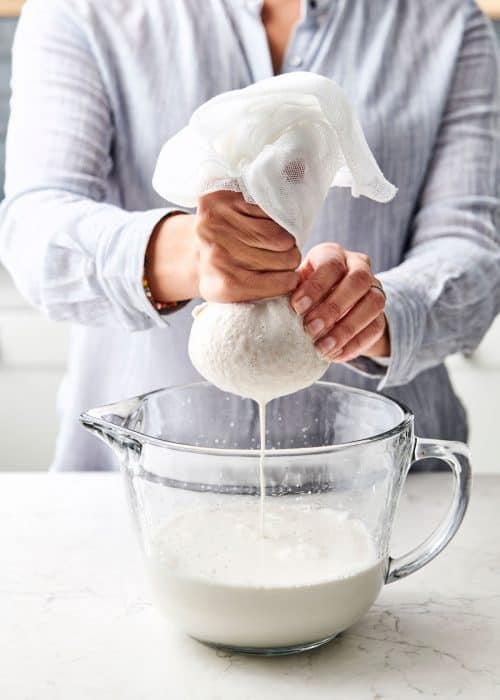 Woman squeezing excess almond milk with a cheese cloth into a large glass bowl in a beautiful white marble kitchen