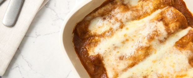 Staub baking dish full of chicken enchiladas & vibrant red sauce and melted cheese on a white marble surface with utensils