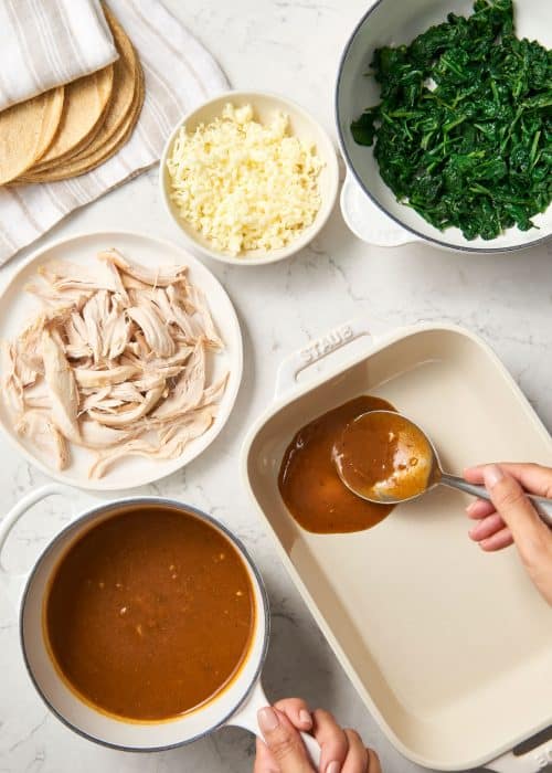 Woman preparing a deep baking dish with mexican red sauce and ingredients for enchiladas on a marble surface