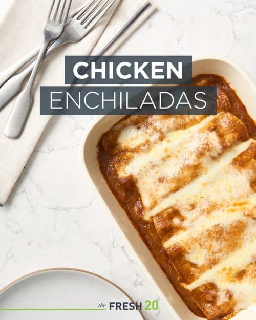 Staub baking dish full of chicken enchiladas & red sauce and melted cheese on a white marble surface with utensils