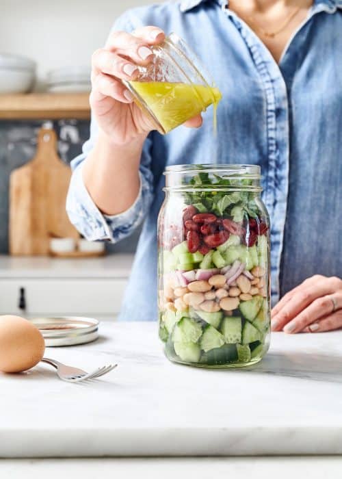 Woman pouring dressing into mason jar salad with hard boiled egg in a white marble kitchen