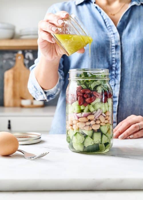 Woman pouring dressing into mason jar salad with hard boiled egg in a white marble kitchen