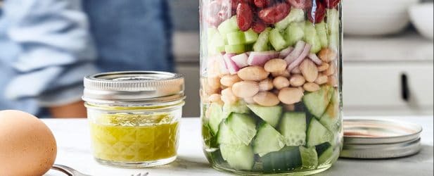 Mason jar filled with vegetable salad next to a mini jar of dressing & a hard boiled egg in a white marble kitchen