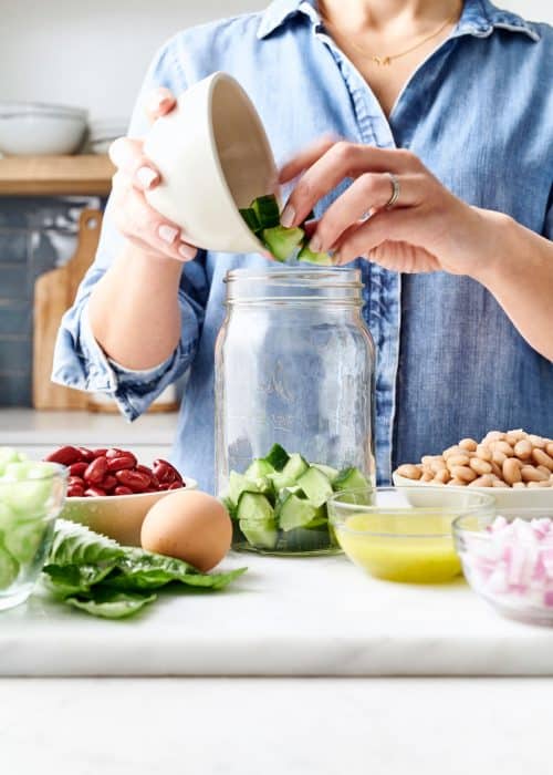 Woman filling glass mason jar with salad & bowls of vegetable ingredients in a white marble kitchen