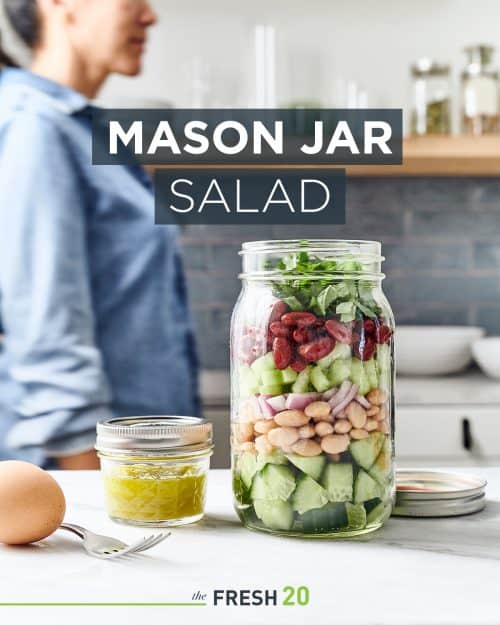 Woman walking by a mason jar filled with salad next to a mini jar of dressing & a hard boiled egg in a white marble kitchen