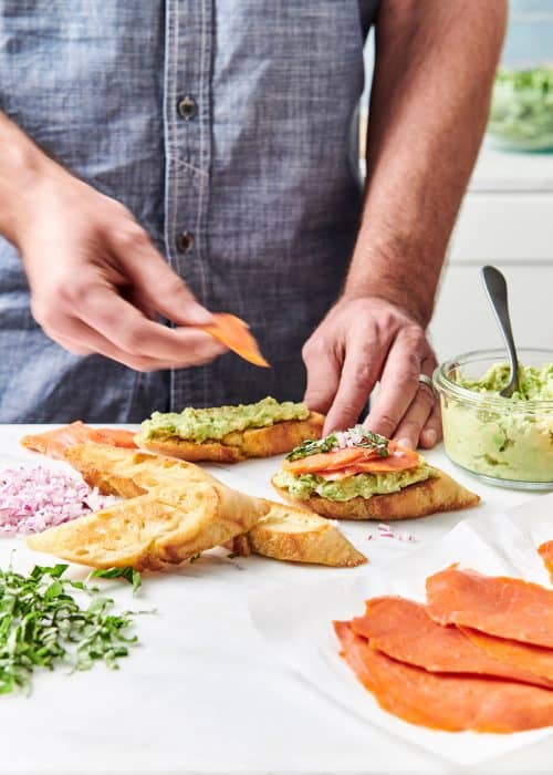 Man preparing smoked salmon avocado toast in a white marble kitchen full of vibrant fresh ingredients