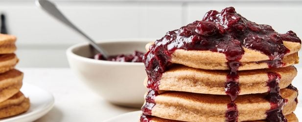 Tall stack of fluffy sweet potato pancakes on a cream plate coated in a thick sweet blueberry compote in a white marble kitchen