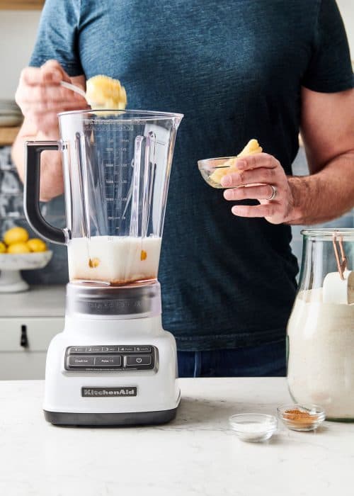 Man making sweet potato pancake batter with spices and flour in a KitchenAid blender on a white marble surface