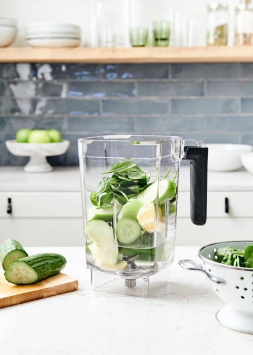 Blender full of fresh cucumber apple ginger green juice fruit & vegetables & a wood cutting board in a beautiful white marble kitchen