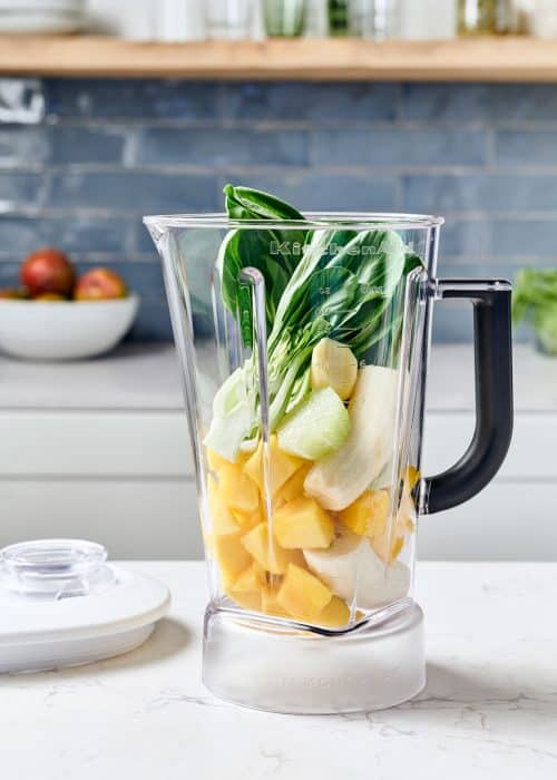 Blender full of fresh mango, bannanas, lime & bok choy in a beautiful white marble kitchen