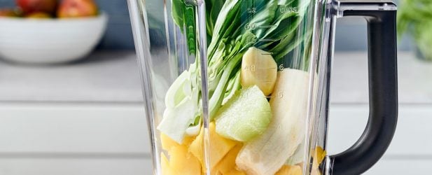 Blender full of fresh mango, bannanas, lime & bok choy in a beautiful white marble kitchen