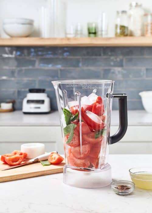 Blender full of tomatoes, basil & red bell peppers next to chopped ingredients on a wood cutting board in a white marble kitchen