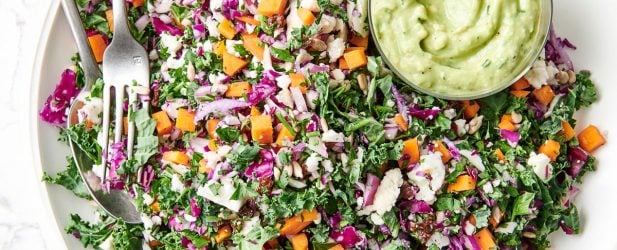 Vibrant vegan chopped salad with a side of avocado dressing on a cream plate with utensils on white marble