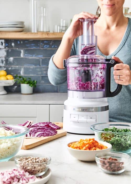 Woman chopping red cabbage in food procesor surrounded with bowls of fresh cut vegetables for a vegan chopped salad in a beautiful white marble kitchen