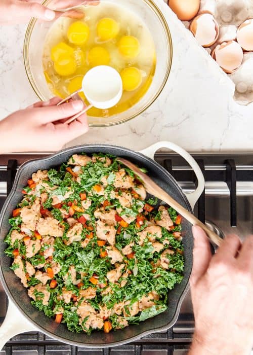 Scooping egg yolks from glass bowl into a skillet full of meat, bell peppers & kale on a cooktop