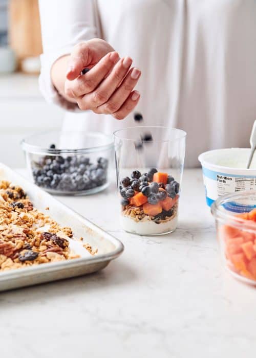 Woman filling a perfect parfait glass with yogurt, blueberries & papaya next to a baking sheet of toasted homemade granola