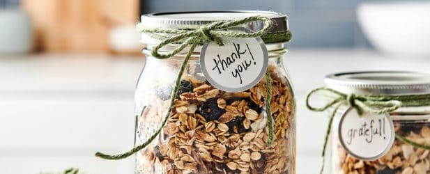 2 mason jars full of decadent homemade DIY granola wrapped with a green twine & a thank you tag on a white marble surface