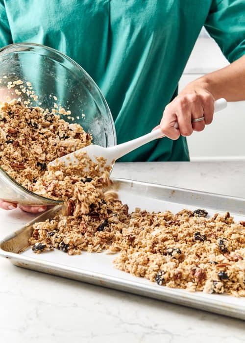 Woman scoops out decadent DIY homemade granola from a glass bowl onto a baking sheet on a white marble countertop