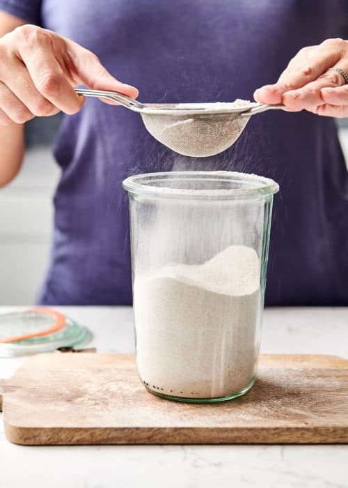 Woman sifting DIY easy decadent waffle mix into a glass containter on a wooden surface