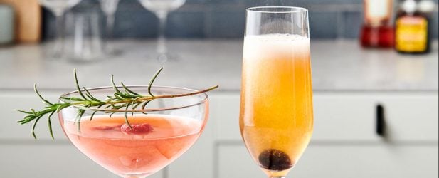 Two glasses filled with vibrant Mother’s Day cocktails garnished with rosemary & berries in a white marble kitchen