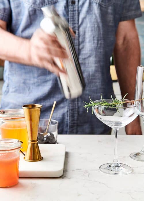 Man making Mother's Day cocktails in a shaker next to a champagne glass garnished with rosemary on a white marble surface